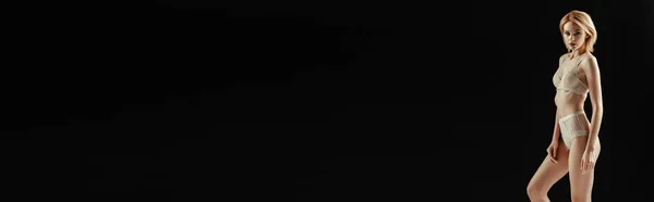 Панорамний знімок привабливої жінки в нижній білизні дивиться на камеру ізольовано на чорному — стокове фото