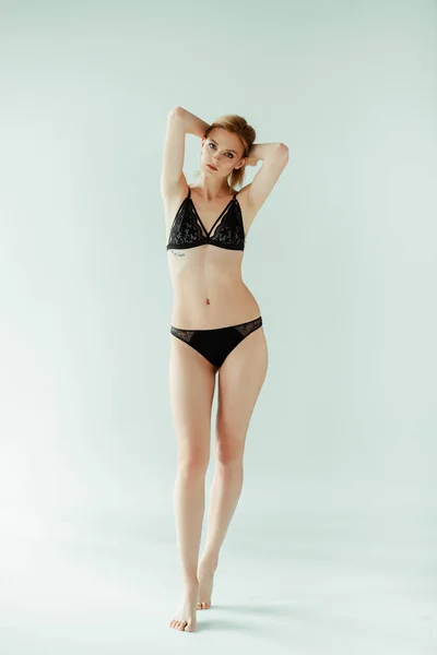 Mulher sexy em lingerie de renda preta com as mãos perto da cabeça olhando para a câmera no fundo cinza — Fotografia de Stock