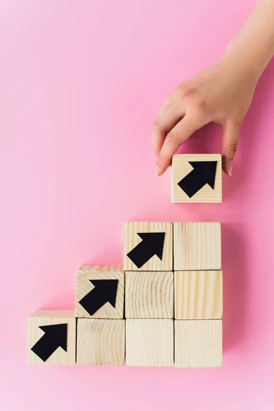 Vista parcial de la mano cerca de bloques de madera con flechas negras sobre fondo rosa, concepto de negocio - foto de stock