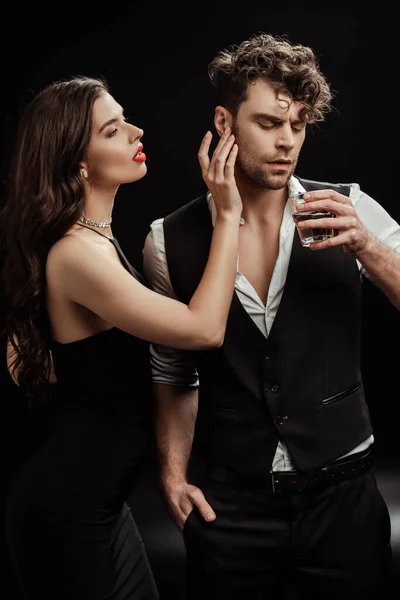 Sexy woman touching boyfriend with glass of whiskey on black — Stock Photo