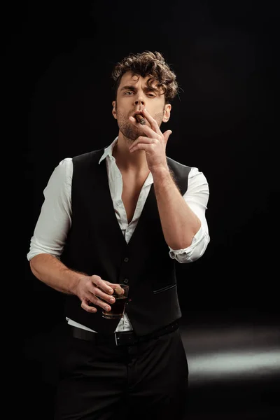 Handsome man looking at camera while smoking cigar and holding glass of whiskey on black background — Stock Photo