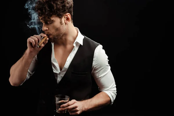 Handsome man smoking cigar and holding glass of whiskey isolated on black — Stock Photo