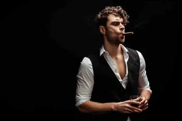 Bel homme en chemise et gilet tenant verre de whisky et cigare fumant isolé sur noir — Photo de stock