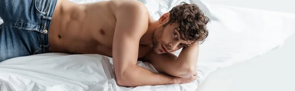 Panoramic shot of handsome muscular man looking at camera on bed isolated on white — Stock Photo