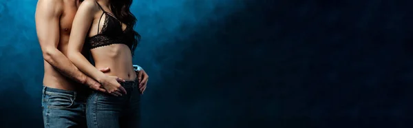 Panoramic crop of muscular man embracing woman in jeans and bra on black background with smoke — Stock Photo