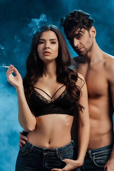 Schöner Mann ohne Hemd umarmt Freundin in BH Zigarette rauchend auf schwarzem Hintergrund mit Rauch — Stockfoto
