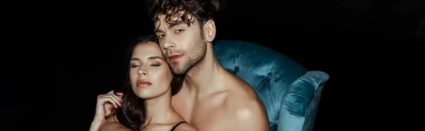 Colpo panoramico di uomo senza maglietta abbracciando fidanzata con gli occhi chiusi sulla poltrona isolata su nero — Foto stock