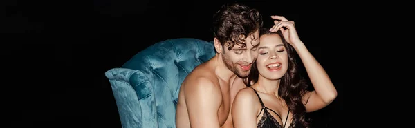 Horizontal image of shirtless man sitting near smiling woman in bra on armchair isolated on black — Stock Photo