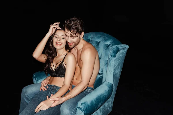 Cheerful shirtless man embracing woman in bra and jeans on armchair isolated on black — Stock Photo