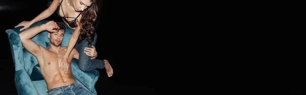 Colheita horizontal de mulher no sutiã tocando tronco muscular de namorado bonito em poltrona isolada em preto — Fotografia de Stock