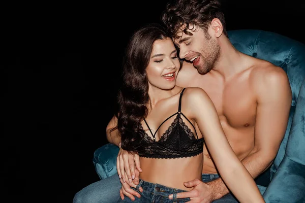 Uomo senza maglietta sorridente mentre abbraccia la donna sexy in reggiseno sulla poltrona isolata sul nero — Foto stock