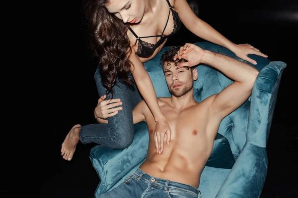 Passionate girl touching torso of boyfriend on armchair isolated on black — Stock Photo