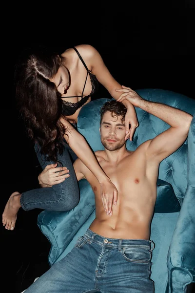 Bonito homem sem camisa tocando mulher em sutiã e jeans em poltrona isolado em preto — Fotografia de Stock