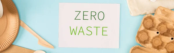 Panoramic crop of card with zero waste lettering near disposable tableware and cardboard egg tray on blue background, ecology concept — Stock Photo
