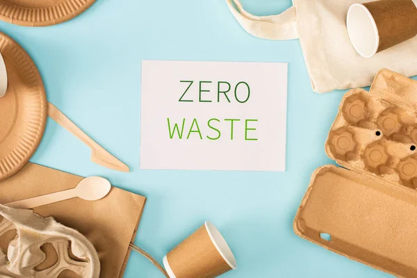 Top view of card with zero waste lettering near disposable tableware and cardboard egg tray on blue background, ecology concept — Stock Photo