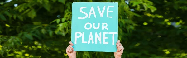 Imagen panorámica del hombre sosteniendo pancarta con salvar nuestro planeta letras con árboles en el fondo, concepto de ecología - foto de stock