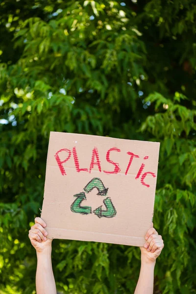 Обрезанный вид человека, держащего плакат с пластиковой надписью и переработанный знак с деревьями на заднем плане, экологическая концепция — стоковое фото