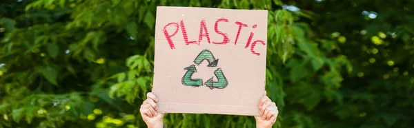 Panoramic crop of man holding placard with recycle sign and plastic lettering with trees at background, ecology concept — Stock Photo