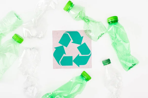 Vista superior de garrafas de plástico amassadas perto do cartão com sinal de reciclagem no fundo branco, conceito de ecologia — Fotografia de Stock