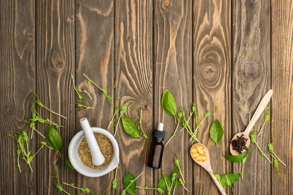 Vue du dessus des pilules dans les cuillères, mortier, herbes vertes et bouteille sur la surface en bois, concept de naturopathie — Photo de stock