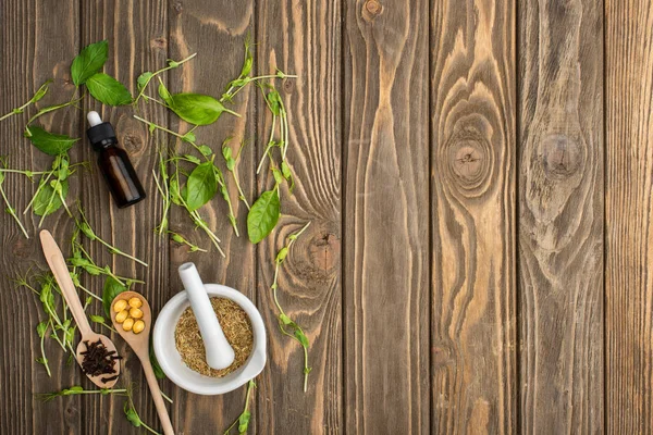 Top view of pills in spoons, mortar, green herbs and bottle on wooden surface, naturopathy concept — Stock Photo