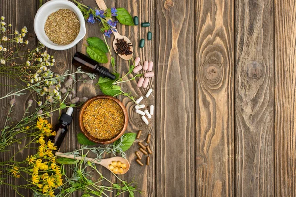 Vista superior de pílulas em colheres, ervas verdes e flores silvestres na superfície de madeira, conceito de naturopatia — Fotografia de Stock