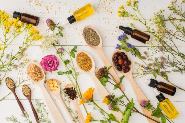 Vista dall'alto di erbe in cucchiai vicino a fiori e bottiglie su sfondo di legno bianco, concetto di naturopatia — Foto stock