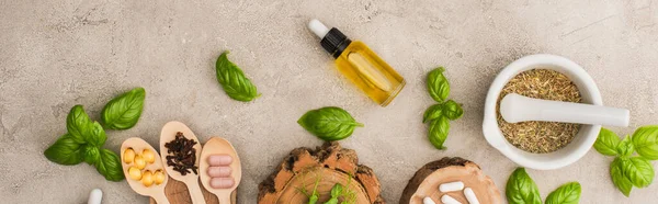 Panoramic shot of herbs, green leaves, mortar with pestle, bottle and pills in wooden spoons on concrete background, naturopathy concept — Stock Photo