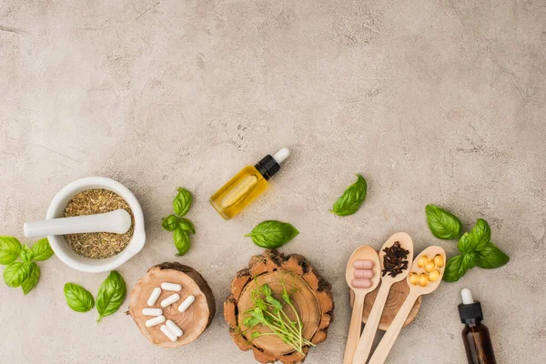 Vista superior de hierbas, hojas verdes, mortero con pestle, botellas y pastillas en cucharas de madera sobre fondo de hormigón, concepto de naturopatía - foto de stock