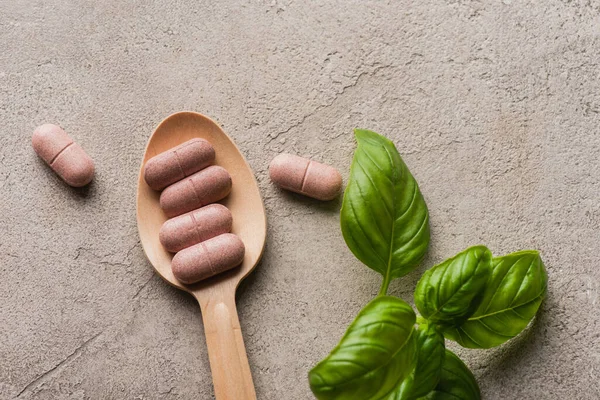 Vista superior de hojas verdes y píldoras en cuchara de madera sobre fondo de hormigón, concepto de naturopatía - foto de stock