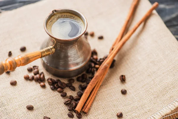 Selektiver Fokus von Cezve mit Kaffee in der Nähe von Zimtstangen und Kaffeebohnen auf Sacktuch — Stockfoto