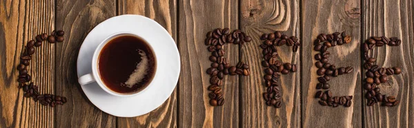 Vista superior de la taza de café en platillo y letras de café hechas de frijoles en la superficie de madera, plano panorámico - foto de stock