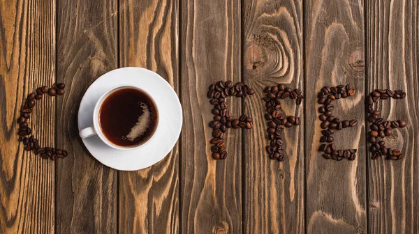 Top view of cup of coffee on saucer and coffee lettering made of beans on wooden surface — Stock Photo