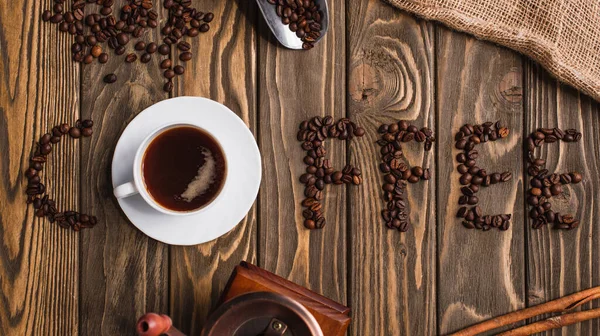 Top view of cup of coffee on saucer and coffee lettering made of beans on wooden surface — Stock Photo