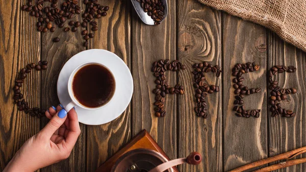 Vista ritagliata della donna che tiene una tazza di caffè sul piattino vicino alla scritta del caffè fatta di fagioli sulla superficie di legno — Foto stock