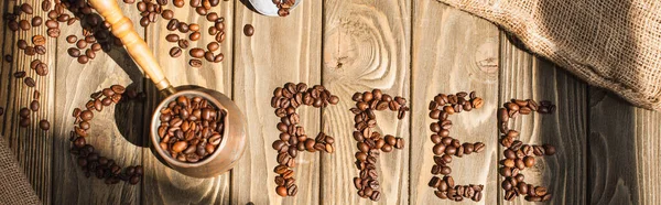 Vista superior de letras de café hechas de frijoles y cezve a la luz del sol en la superficie de madera, plano panorámico - foto de stock