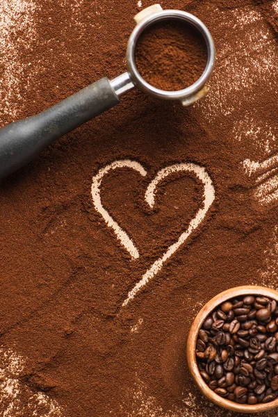 Vista superior del corazón dibujado en el café molido cerca de frijoles en tazón y portafilter - foto de stock