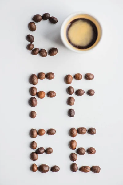 Top view of coffee lettering made of beans near cup on white surface — Stock Photo