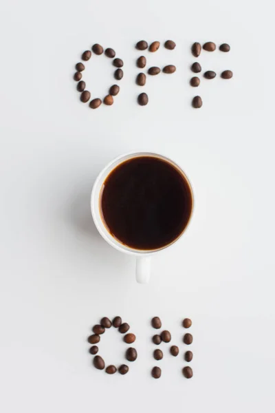 Draufsicht auf ein und aus Schriftzug aus Kaffeebohnen nahe Tasse auf weißer Oberfläche — Stockfoto