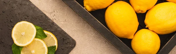 Top view of whole yellow lemons in box and sliced on board with mint green leaves on beige concrete surface, panoramic shot — Stock Photo