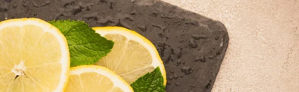 Draufsicht auf mintgrüne Blätter, Zitronenscheiben auf schwarzem Brett auf beiger Betonoberfläche, Panoramaaufnahme — Stockfoto