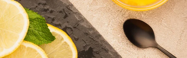 Vue de dessus des feuilles vert menthe, tranches de citron sur le tableau noir près de cuillère sur la surface en béton beige, vue panoramique — Photo de stock