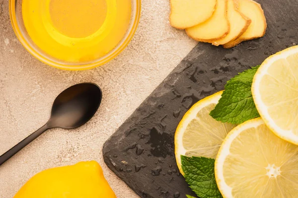 Vue de dessus des feuilles de menthe verte, des tranches de racine de gingembre et de citron et du miel sur le panneau noir sur la surface en béton beige — Photo de stock