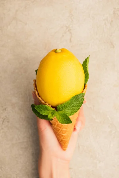 Vista ritagliata di donna che tiene cono cialda con limone giallo con foglie verde menta su superficie di cemento beige — Foto stock