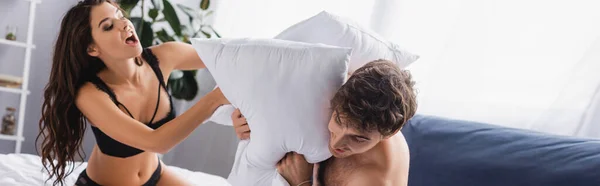 Tiro panorâmico de homem sem camisa e mulher jovem em roupa interior jogando travesseiro jogo de luta na cama — Fotografia de Stock