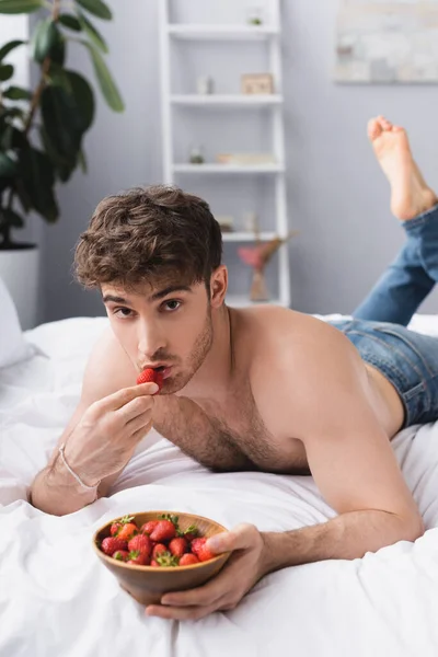 Selective focus of shirtless man lying on bed and holding bowl with ripe strawberries — Stock Photo