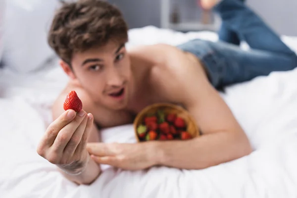 Selektiver Fokus des hemdlosen Mannes, der auf dem Bett liegt und rote Erdbeere in der Hand hält — Stockfoto