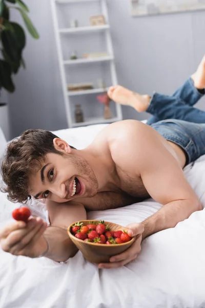 Selektiver Fokus des muskulösen Mannes, der auf dem Bett liegt und rote Erdbeeren in der Schüssel hält — Stockfoto