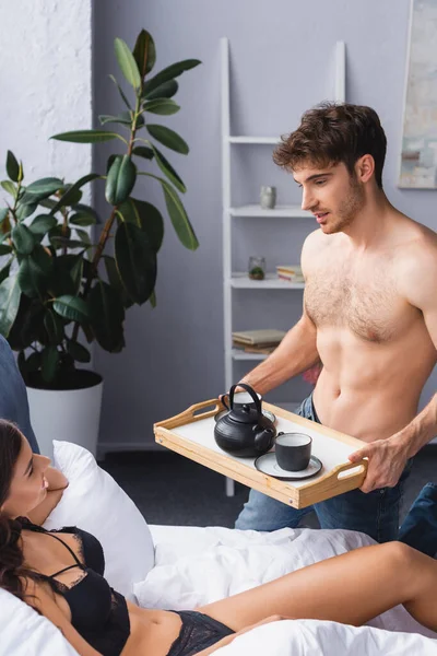 Muskulöser Mann hält Tablett mit Teekanne und Tassen neben verführerischer Frau — Stockfoto