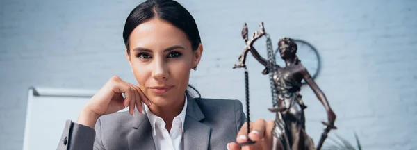 Panoramic shot of brunette lawyer in formal wear touching statuette of justice in office — Stock Photo